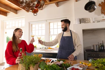 Tuinposter Happy caucasian couple preparing food in kitchen, making a toast with glasses of white wine © wavebreak3