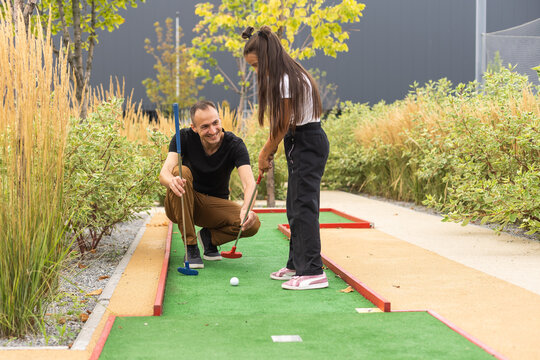 Sharing with golf experience. Cheerful young man teaching his daughter to play mini golf at the day time. Concept of friendly family