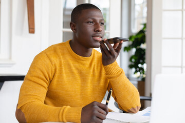 African american man at home writing notes and talking on smartphone