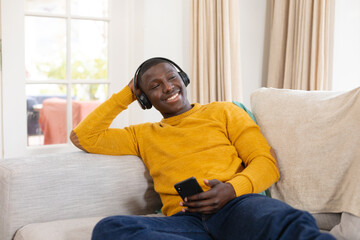 Smiling african american man at home sitting on couch wearing headphones and looking at smartphone