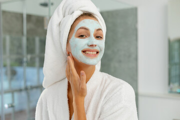 Portrait of happy caucasian woman wearing robe, applying beauty face mask in bathroom