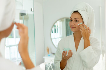 Happy caucasian woman wearing robe, applying beauty face mask on face in bathroom