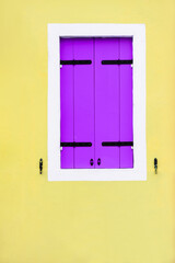 Yellow painted facade of the house and window with lavender shutters.