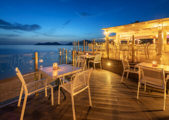 Beautiful cafe on sea coast at night in summer. Landscape with chairs and tables, lamps, lights,...