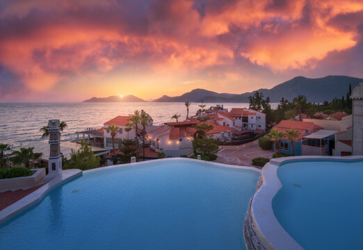 Beautiful swimming pool, sky with pink clouds at colorful sunset. Blue water, sea coast, architecture, palm trees in summer. Luxury resort. Oludeniz, Turkey. Landscape with pool, houses, orange roofs © den-belitsky