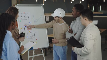 Multi ethnic people in formal wear listening report of qualified male builder in office. Focused engineer in white helmet analysing house model on flip chart.