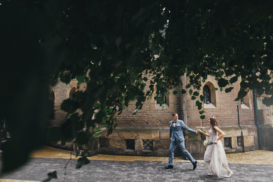 Stylish happy bride and groom running and smiling on background of church under tree leaves. Provence wedding. Beautiful emotional wedding couple in european city.