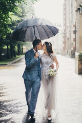 Stylish bride and groom walking under umbrella and kissing on background of old church in rain....
