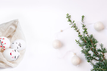 Obraz premium Concept of combination of spring and winter holidays. Christmas garland on tree branch and Easter painted eggs in eco-friendly mesh bags on white table. Handmade workshop. Copy space. Selective focus