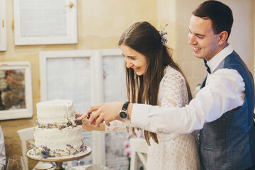 Stylish happy wedding couple cutting together modern cake with lavender in stylish restaurant. Provence wedding reception. Beautiful bride and groom celebrating with delicious wedding cake
