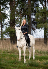 young beautiful blond smiling woman with long hair in black dress riding a white horse with blue eyes in autumn field	