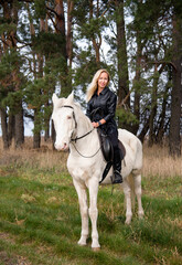 young beautiful blond smiling woman with long hair in black dress riding a white horse with blue eyes in autumn field	