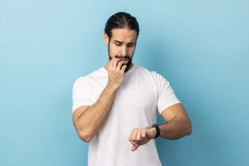 Portrait of man with beard wearing white T-shirt pointing finger on his wrist watch with nervous face, time is out, biting finger nails. Indoor studio shot isolated on blue background.