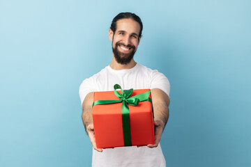 Portrait of handsome delighted man with beard wearing white T-shirt giving wrapped gift box and smiling at camera, happy holiday. Indoor studio shot isolated on blue background.
