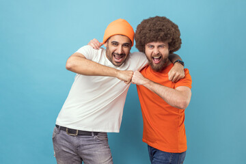 Fototapeta premium Yes we did. Portrait of two handsome happy excited young adult hipster men celebrating triumph together, showing bro fist, power five sign. Indoor studio shot isolated on blue background.