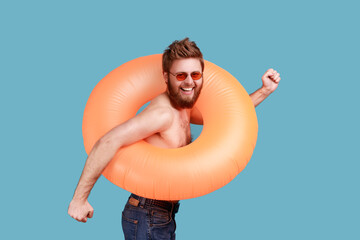 Portrait of excited happy bearded man standing with bright orange rubber ring, looking at camera with toothy smile, enjoying his vacation. Indoor studio shot isolated on blue background.