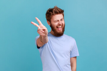 Portrait of happy optimistic bearded man doing victory gesture and winking playfully to camera,...