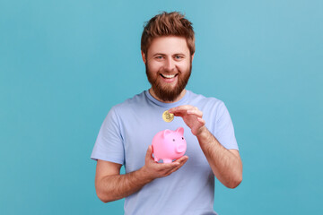 Portrait of happy smiling bearded man looking at camera, putting coin into piggybank, savings of...