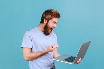 Portrait of handsome astonished surprised bearded man pointing finger at laptop display with big eyes shocked with content. Indoor studio shot isolated on blue background.