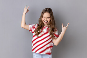 Portrait of little girl wearing striped T-shirt showing rock and roll gesture heavy metal sign,...
