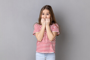 Portrait of dark haired nervous adorable little girl wearing striped T-shirt biting his fingers...