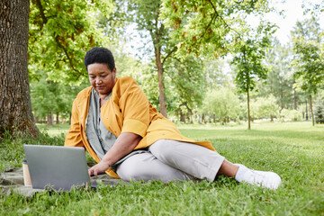 Full length portrait of senior black woman using laptop outdoors while enjoying picnic on green grass in park, copy space