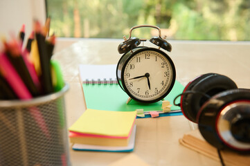 Still life. Focus on a black alarm clock on open notepad with school supplies, audio headset and stationery on blurred foreground. Back to school. Education. Teacher's day concept