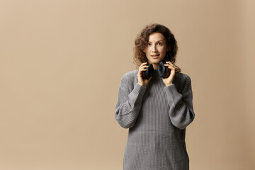 Obraz premium Cheerful enjoyed pretty curly beautiful female in gray casual sweater wear headphones looks at camera posing isolated on over beige pastel background. Sound streaming platform concept. Copy space