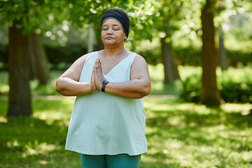 Waist up portrait of mature black woman doing yoga outdoors in green park and holding hands...