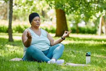 Full length portrait of overweight black woman doing yoga outdoors and meditating with eyes closed on green grass - obrazy, fototapety, plakaty