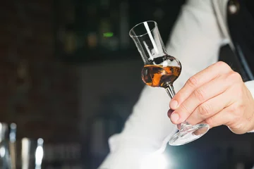Foto op Plexiglas Strong alcohol. A professional bartender in a white shirt and black apron holds a tasting glass, the trend drinks concept. © Oleksandr Slastin