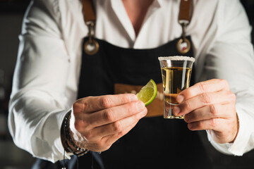 Tequila. A professional bartender in a white shirt and black apron holds a glass of tequila shot, the trend drinks concept.