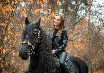 young beautiful smiling woman in black gothic dress riding black Friesian horse  in autumn forest with yellow leaves