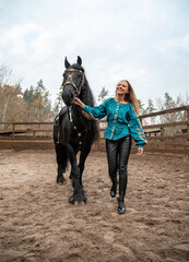 Obraz premium young beautiful smiling woman in ukrainian green embroidery walking with black Friesian horse on a farm in autumn