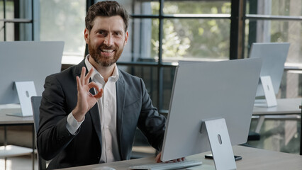 Happy success cheerful bearded 40s Caucasian adult man 30s businessman manager worker working with computer in office smile looking into camera with sign ok good idea done job approve agree advertise