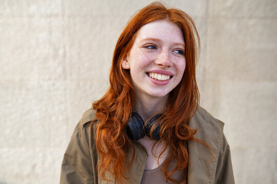 Happy teen stylish cool redhead fashion girl model standing on urban wall background laughing. Close up portrait of pretty joyful smiling teenage hipster girl with red hair looking away outdoors.