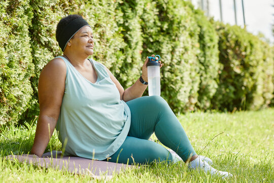 Full length side view of overweight black woman relaxing on grass while working out outdoors with water bottle, copy space