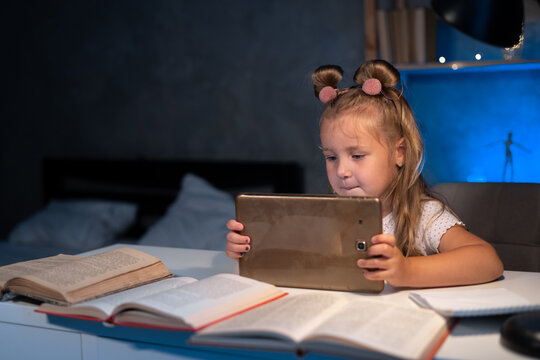 Happy Child Little Girl Using Tablet Computer Sitting At The Table Doing Homework At Night. Elementary School Student Studying Virtually.