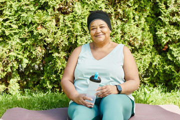 Front view portrait of mature black woman looking at camera and smiling while drinking water at break in outdoor workout - Powered by Adobe