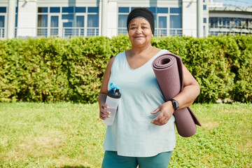 Waist up portrait of mature black woman holding yoga mat outdoors and smiling at camera, copy space