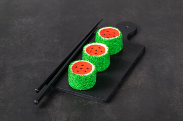 Obraz na płótnie Canvas Children Mousse cream dessert with jelly watermelon in the form of round sweet Japanese sushi, sprinkled with green coconut chips, on a serving board. Dark gray background