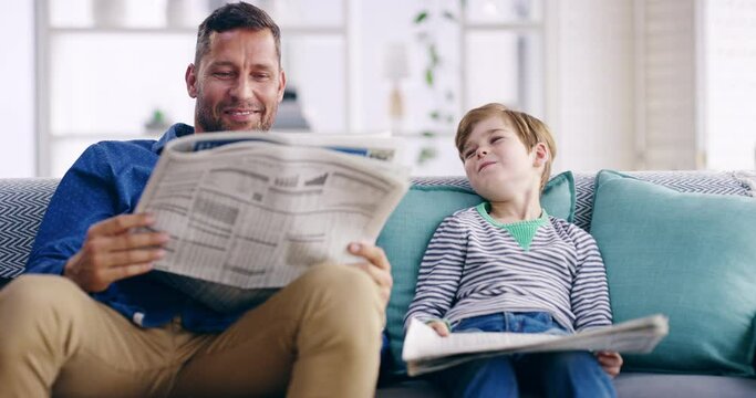 Father and son bonding, reading newspaper and relaxing on a sofa at home. Happy parent smiling, teaching his child the importance of keeping up with current news, talking, laughing and carefree