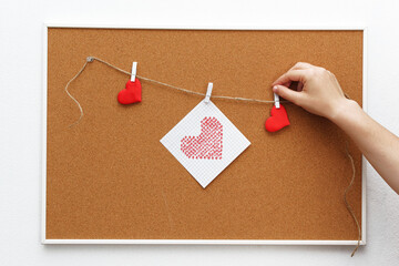 A woman's hand attaches a clothespin with a red heart drawn on a piece of paper to a cork board, space for text.