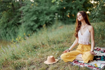 A young hippie woman meditates in nature in the park, sitting in a lotus position on her colorful plaid and enjoying harmony with the world in eco-clothing