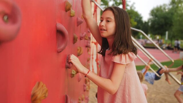 A little girl climbs a children's climbing wall on a playground in the park.