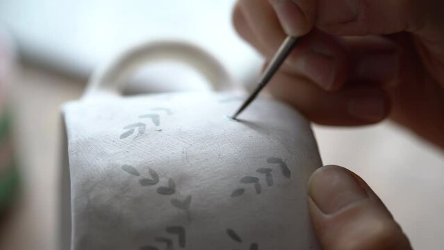Hands of professional artisan hold white ceramic mug drawing pattern with dark paint using brush on blurred background. Woman enjoys working with handmade pottery craft in studio extreme closeup