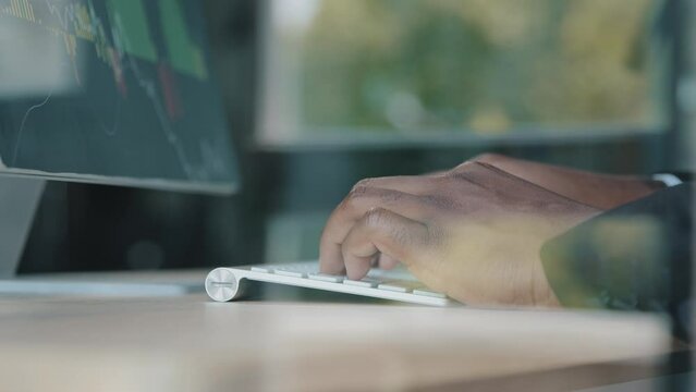 Close up behind glass view of unknown african businessman trader programmer IT specialist work on computer in monitoring control room typing on keyboard programming trading online developing software