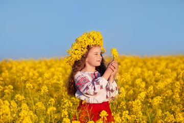 Naklejka na ściany i meble Ukrainian child girl in traditional embroidery and yellow wreath in field of yellow flowers against blue sky. Pray for Ukraine. Ukraine's Independence Flag Day. Constitution day. Symbols of Ukraine.