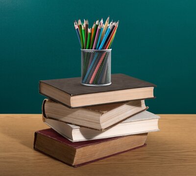 Stack of books and holder with stationery on table near chalkboard in classroom