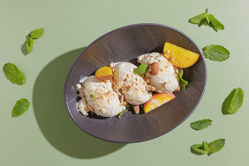 three scoops of ice cream with nuts and peach slices on a dark plate on a green background with a shadow from a palm tree, top view space for text